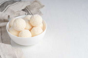 Fototapeta na wymiar Coconut organic truffles dessert or homemade nut vegan energy balls served in bowl with textile towel on white wooden background at kitchen ready to eating. Image with copy space, horizontal