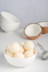 Coconut organic sweet truffles or homemade nut vegetarian energy balls with filling of shredded coconut and curd cheese served in bowl on white wooden table at kitchen against gray wall. Vertical