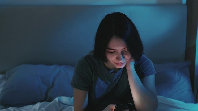 Night online. Bedtime insomnia. Social media addiction. Bored relaxed woman using mobile phone surfing Internet sitting alone in bed late in dark home bedroom with blue light.