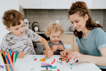 Fototapeta premium A young pretty mother is sitting at the kitchen table with her preschool-aged daughter and school-aged son making something out of plasticine. The mother is spending free time with her children.