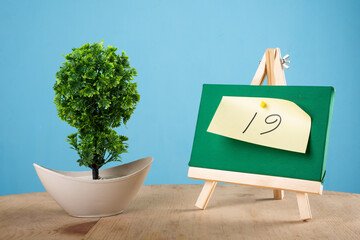  19 written on a sticky note on a mini green notice board with a plant with blue background.