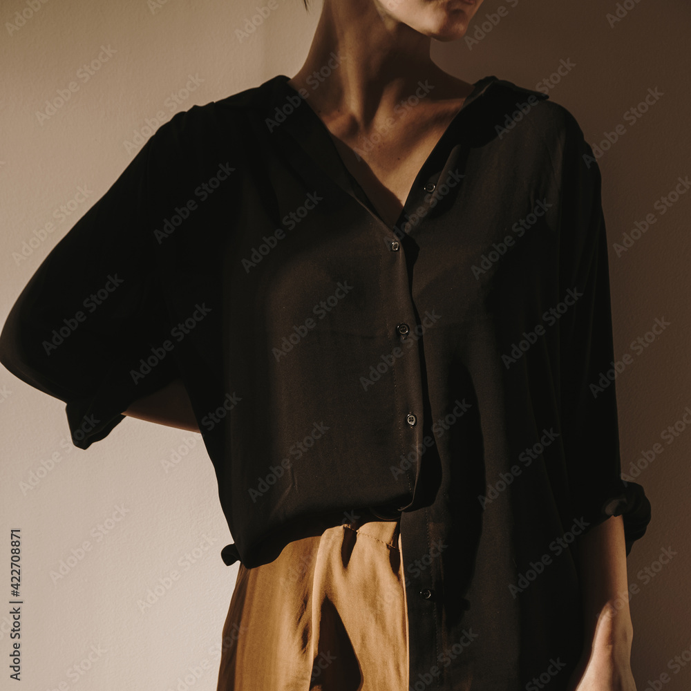 Wall mural Young beautiful woman in black shirt and brown trousers staying against the wall. Tan sunlight shadows on the wall. Minimal aesthetic beauty, fashion concept for magazine, blog, social media.