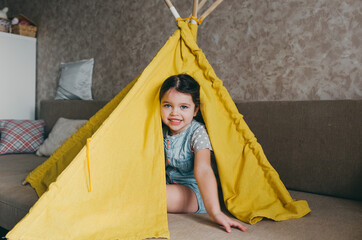 a little girl sits and smiles inside a yellow teepee. home games and entertainment for children © Tatsiana