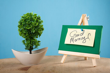 Good morning written on a sticky note on a mini green notice board with a plant with blue background
