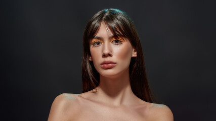 Portrait of naked young brunette woman with big brown eyes and colorful makeup posing for camera isolated over black background