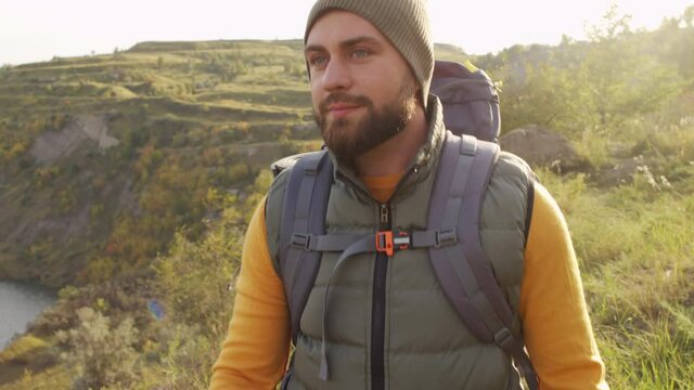 Slowmo tracking of bearded male tourist with backpack hiking on hill near quarry lake on sunny day