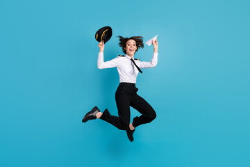 Full length photo of charming cheerful young woman jump up air hold plane origami hat isolated on blue color background