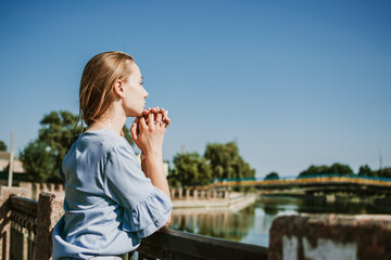 Lonely pensive Young blonde woman in blue dress standing near the city river in summer sun day. Alone deep in thought woman outdoors