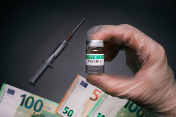 Hand holds COVID-19 vaccine, with money on the background. Medical syringe and Covid-19 vaccine with cash. Cost of coronavirus vaccine. 