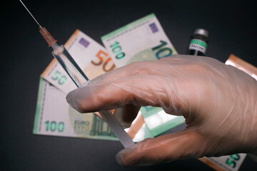 Hand holds syringe, cash and COVID-19 vaccine on the background. Medical syringe with Covid-19...