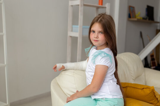 Child With A Cast On A Broken Wrist Or Arm Smiling And Having Fun On A Couch. Positive Attitude, Recovery And Kid Concept.