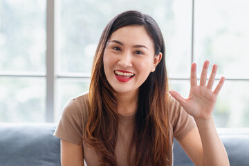 Close up portrait head shot of Asian long black and brown hair young beautiful female wearing brown shirt sitting on blue sofa smiling hold hand up to say hi happily in front glass windows background