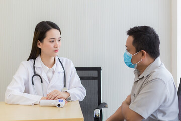 Asian beautiful young woman doctor talking with a man patient about his pain and symptom while they put on a face mask to prevent Coronavirus disease and Thermometer on table in hospital. 