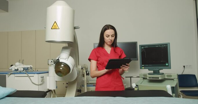 Close up portrait of young caucasian woman doctor in red uniform, holding digital tablet pc, posing near modern ultrasonic lithotripter machine in urology medical center