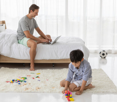 Father Sitting On Bed And Working With Notebook Computer While His Mixed-race Son Playing Colorful Wooden Block In Home Bedroom. Idea For Lifestyle Of Working From Hom