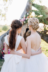 two brides with bouquets stand with their backs against the background of a decorated tree in a meadow