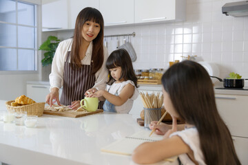 A cute little 7 years old Asian girl sitting and doing homework in kitchen while her mother...