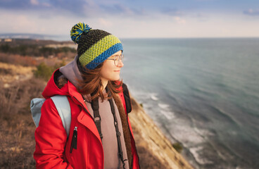 Fototapeta na wymiar Young happy woman in cap and red jacket traveling enjoying fresh air on the rocky seashore, solo travel