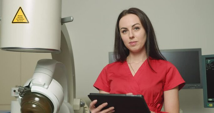Close up portrait of young caucasian woman doctor in red uniform, holding digital tablet pc, posing near modern ultrasonic lithotripter machine in urology medical center