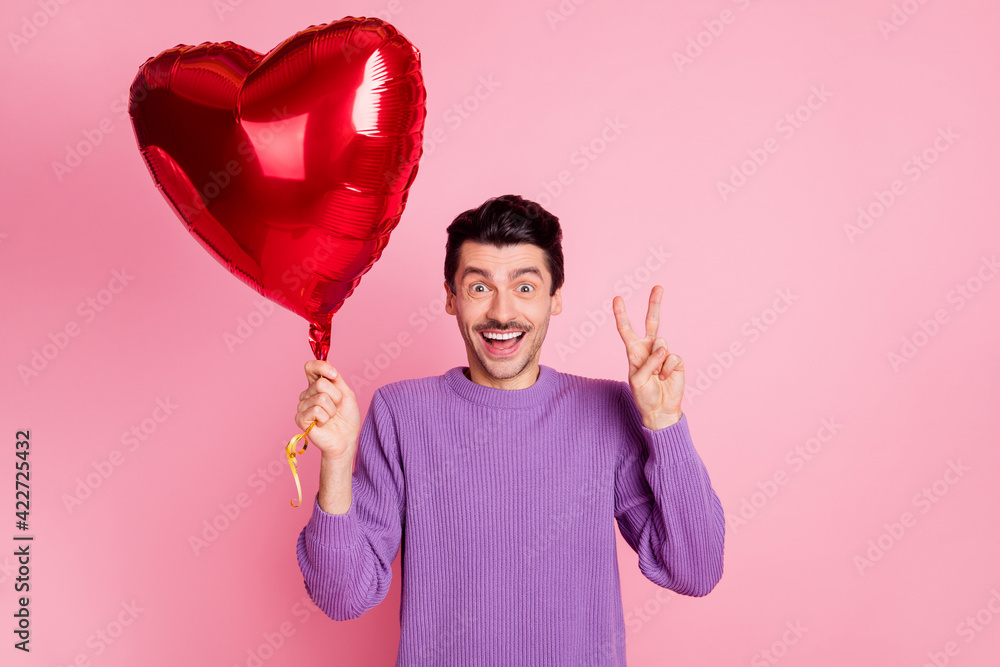 Poster portrait of attractive cheerful guy holding heart air ball showing v-sign isolated over pink pastel 