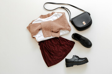 Folded clothes for women fashion urban basic outfit. Female spring look autumn outfit burgundy skirt beige sweater black shoes bag white tshirt on white background. school uniform top view.