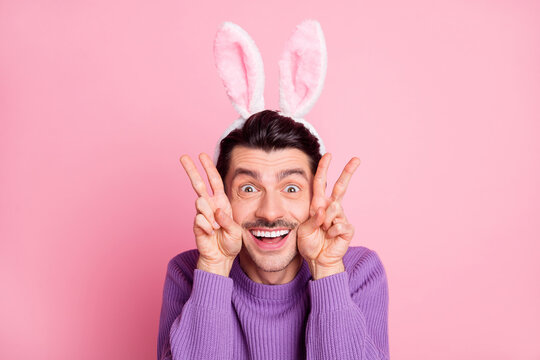 Photo of charming funky young man dressed purple pullover rabbit ears showing v-signs cheeks isolated pink color background