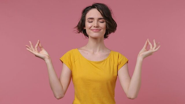 Brunette short hairdo young woman 20s in basic casual yellow t-shirt hold spreading hands in yoga om gesture relaxing meditating trying to calm down isolated on pastel pink background studio portrait