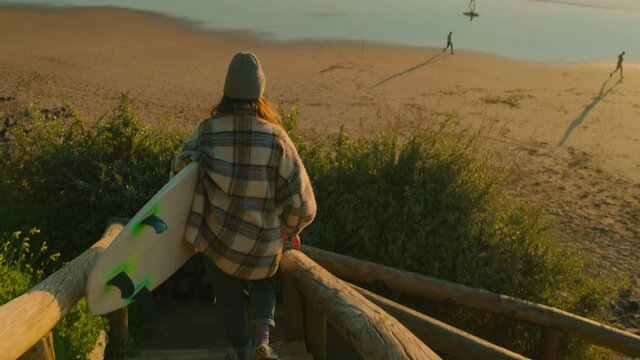 Handheld shot of camera follow young woman in casual clothes walk down wooden stairs to secluded beautiful beach for evening surfing session. Millennial female surfer or vanlife nomad