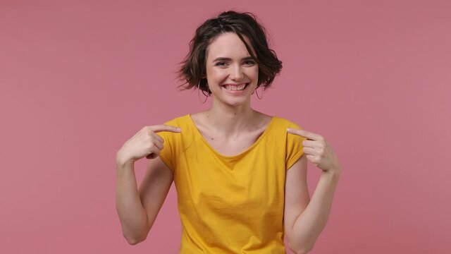 Confident brunette short hairdo young woman 20s wears basic casual yellow t-shirt looking camera charming smile pointing fingers on herself blinking isolated on pastel pink background studio portrait
