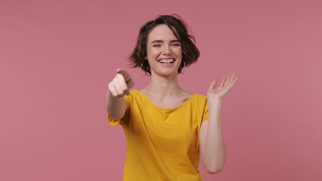 Excited cheerful fun short hairdo young woman 20s wears basic casual yellow t-shirt look camera laugh smiling watch comedy movie pointing index finger on you isolated on pastel pink background studio