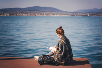 Fototapeta premium A young woman artist sitting on a pier on a sunny day and draws a landscape in a sketchbook