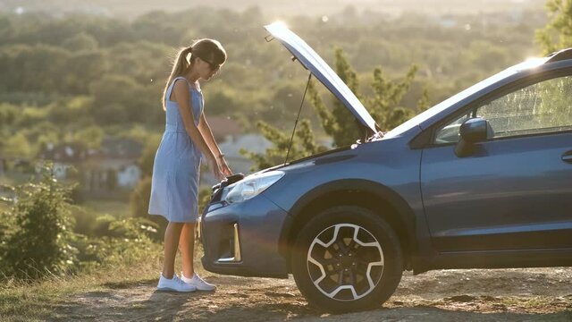 Young female driver standing near a broken car with open up hood inspecting her vehicle engine and waiting for help.