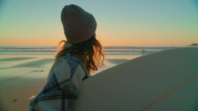 Handheld cinematic shot of urban millennial young woman walk with surfboard under arm on epic beautiful beach at sunset, after evening session of surfing. California vibes lifestyle