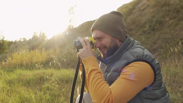Slowmo side view shot of happy bearded man in puffy vest and beanie hat smiling and taking photos outside on sunny day