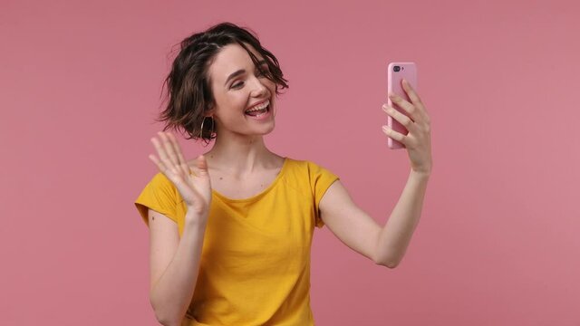 Young woman 20s wears yellow t-shirt get video call using mobile cell phone doing selfie videoconference talk conducting pleasant conversation greet with hand isolated on pastel pink background studio