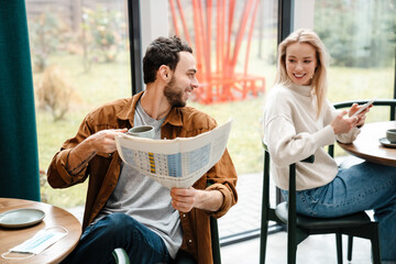 Smiling man and woman using cellphone and reading newspaper