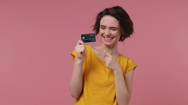 Brunette fun short hairdo young woman 20s wear basic casual yellow t-shirt pointing index finger on mockup plastic credit bank card showing thumbs up isolated on pastel pink background studio portrait