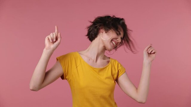 Short hairdo young woman 20s in yellow t-shirt dance clenching fists waving rising expressive gesticulate hands fool around have fun celebrate play fluttering hair isolated on pastel pink background