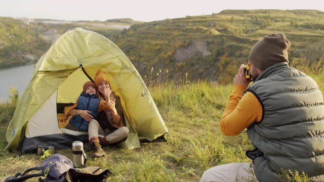 Medium shot of young man using photo camera and taking pictures of happy young woman and 5-year-old boy sitting in tent pitched on green hill overlooking lake