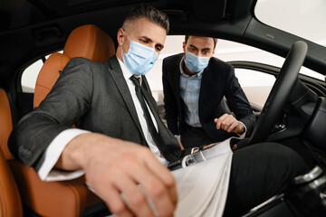 Mid white manager in face mask showing car to customer in showroom