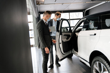 Mid white manager in face mask showing car to customer in showroom