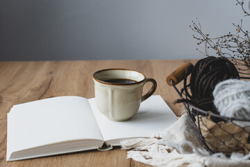 Eine rustikale Kaffeetasse auf einem offenen Buch und Strickzeug in einem Korb auf einem braunen...