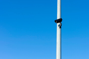 Black CCTV camera on gray pole against blue sky. Sunny winter day. Side view.