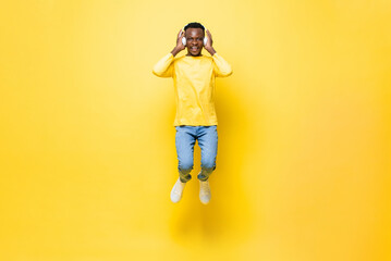 Fototapeta na wymiar Happy young African man wearing headphones listening to music and jumping in yellow isolated studio background