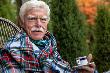 An older man wrapped in a blanket relaxes on a porch in the garden, drinking coffee.