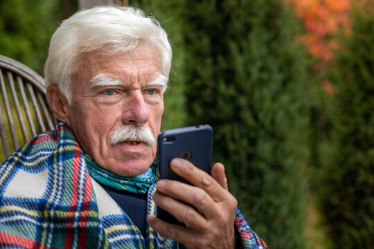 An Elderly Gentleman Is Having A Phone Call. Senior Man Sitting In The Rocking Chair During His Smartphone Conversation. Mature Man Using His Cellular On The Veranda.