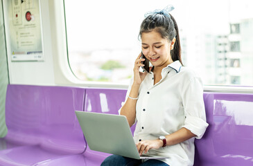 Asian businesswoman talking on cellphone and working on laptop while traveling by sky train. Business travel concept.