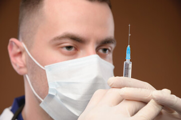 A young male Caucasian doctor in a mask holds a syringe with an injection with his gloved hand. Vaccines and vaccinations and injections concept. Focus on the syringe