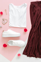 Female spring look summer outfit skirt shoes sneakers basic tshirt bag. Folded clothes for women fashion urban basic outfit with accessories flowers make up cosmetics on pink background. Top view