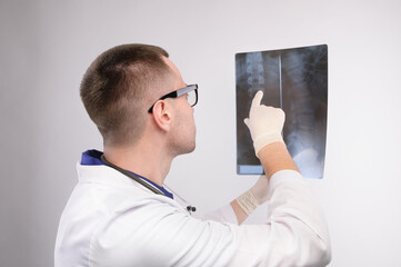 A young male doctor in glasses with a stethoscope around his neck holds an X-ray image of the spine...